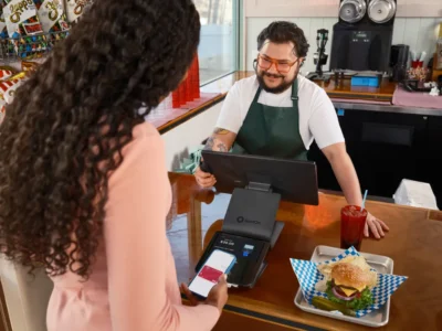 Dono de lanchonete atende cliente que paga por aproximação com o celular, representando o microempreendedor individual